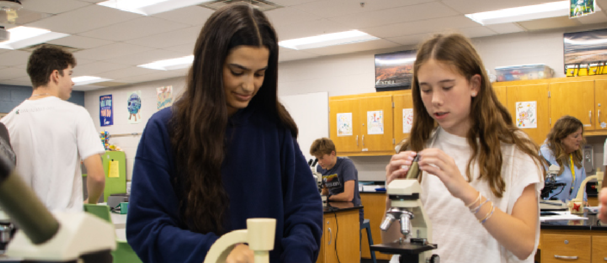 MENTORSHIP MAGNIFIED:  Sophomore Zayna Hafeez, Teacher Assistant (left), helps freshman Abby Brannon (right)  through a microscope lab, showing how to adjust the magnification. By helping prepare and position the slides, Hafeez assisted students in seeing differences between cells.